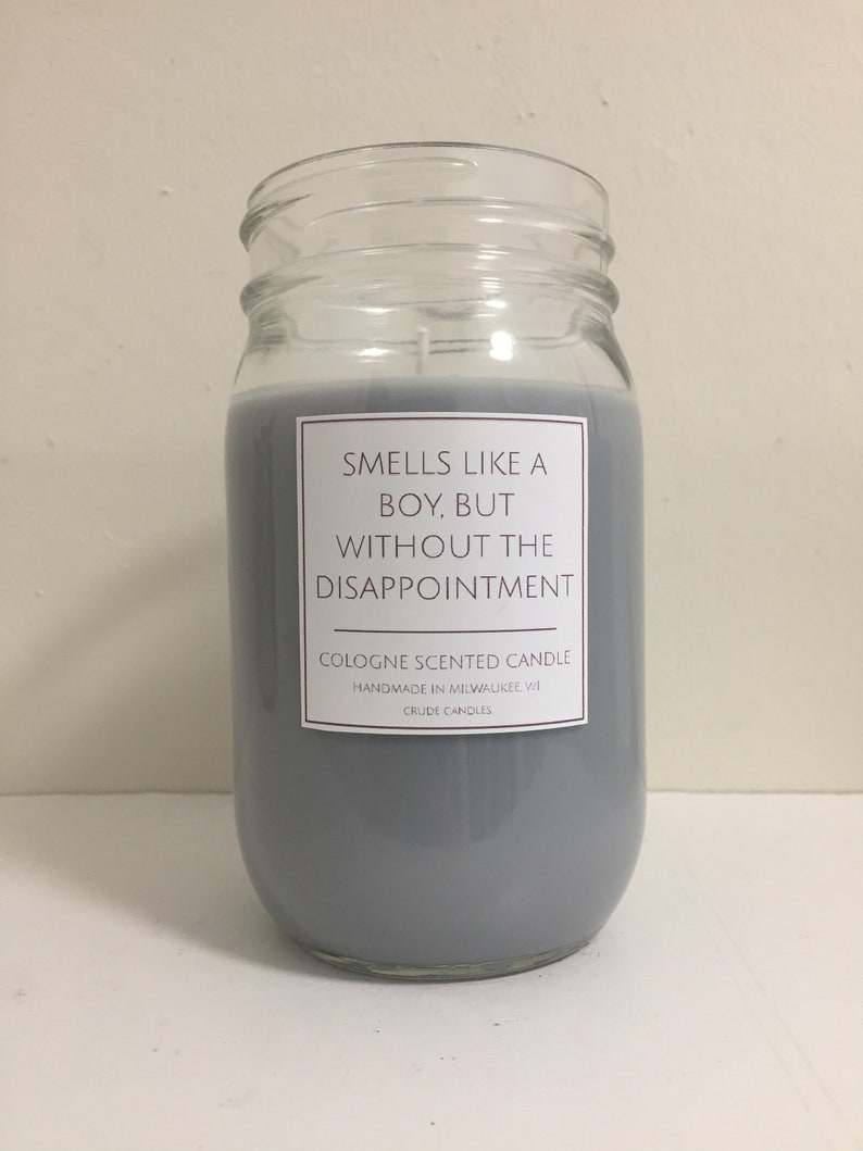 Puede incluir: Una vela de vidrio gris en un frasco con una etiqueta blanca que dice "Huele como un chico, pero sin la decepci&oacute;n." La etiqueta tambi&eacute;n dice "Vela perfumada con colonia" y "Hecho a mano en Milwaukee WI".