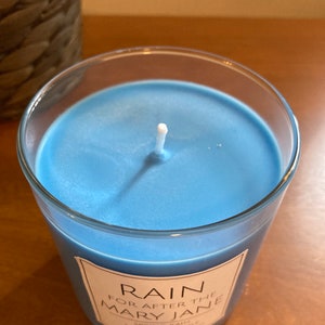 K&ouml;nnte beinhalten: Ein blaues Glas-Kerzengef&auml;&szlig; mit einem wei&szlig;en Docht und einem wei&szlig;en Etikett mit der Aufschrift "RAIN FOR AFTER THE MARY JANE SPRING RAIN SCENTED CANDLE HAND POURED & WAXED"