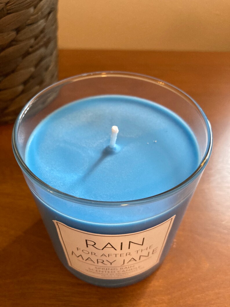 K&ouml;nnte beinhalten: Ein blaues Glas-Kerzengef&auml;&szlig; mit einem wei&szlig;en Docht und einem wei&szlig;en Etikett mit der Aufschrift "RAIN FOR AFTER THE MARY JANE SPRING RAIN SCENTED CANDLE HAND POURED & WAXED"