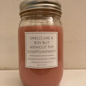Puede incluir: Una vela de vidrio con cera rosa y una etiqueta blanca que dice "Huele como un chico, pero sin la decepci&oacute;n." La etiqueta tambi&eacute;n dice "Vela perfumada con colonia", "Hecho a mano en Milwaukee, WI" y "Crude Candles".