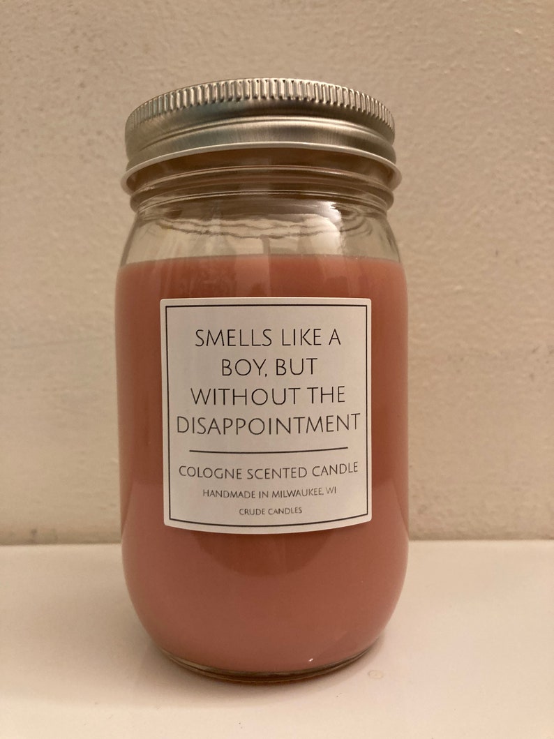 Puede incluir: Una vela de vidrio con cera rosa y una etiqueta blanca que dice "Huele como un chico, pero sin la decepci&oacute;n." La etiqueta tambi&eacute;n dice "Vela perfumada con colonia", "Hecho a mano en Milwaukee, WI" y "Crude Candles".