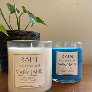 K&ouml;nnte beinhalten: Zwei Glaskerzenbeh&auml;lter mit wei&szlig;em und blauem Wachs. Die Beh&auml;lter tragen Etiketten mit der Aufschrift "RAIN FOR AFTER THE MARY JANE" und "SPRING RAIN SCENTED CANDLE HANDMADE IN MILWAUKEE, WI CRUDE CANDLES".