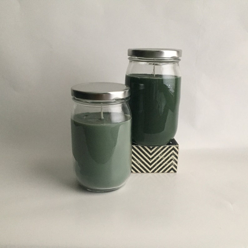 Puede incluir: Dos tarros de vidrio con tapas de metal llenos de velas arom&aacute;ticas verdes. Los tarros est&aacute;n sobre una superficie con un patr&oacute;n blanco y negro.