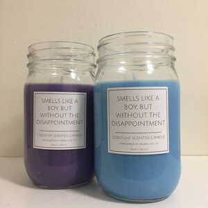 Puede incluir: Dos velas de vidrio en tarros con cera morada y azul. Las etiquetas en los tarros dicen "Smells like a boy, but without the disappointment. Cologne scented candle. Handmade in Milwaukee, WI. Crude Candles."