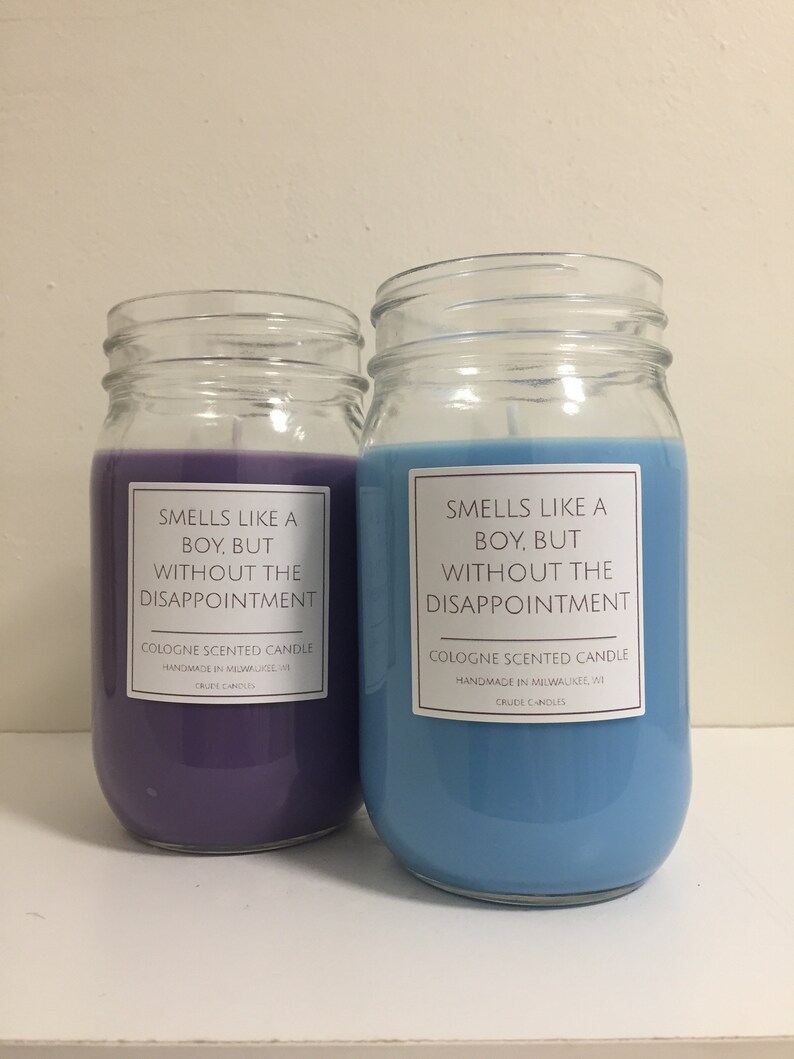 Puede incluir: Dos velas de vidrio en tarros con cera morada y azul. Las etiquetas en los tarros dicen "Smells like a boy, but without the disappointment. Cologne scented candle. Handmade in Milwaukee, WI. Crude Candles."