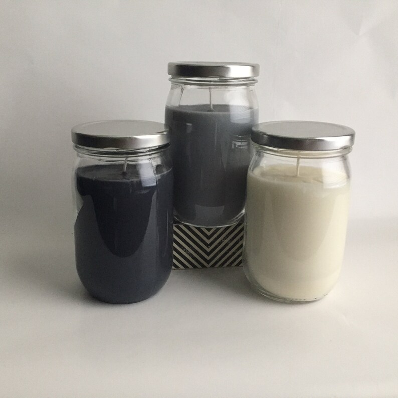 Puede incluir: Tres tarros de vidrio para velas con tapas plateadas. Las velas son en tonos grises y blancos.