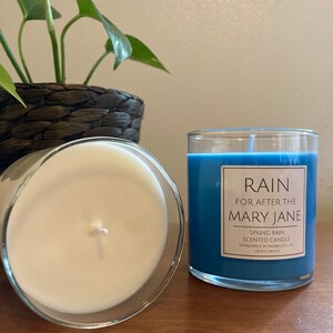 K&ouml;nnte beinhalten: Zwei Kerzen, eine mit blauem Wachs und die andere mit wei&szlig;em Wachs. Die blaue Kerze hat ein Etikett mit der Aufschrift "RAIN FOR AFTER THE MARY JANE SPRING RAIN SCENTED CANDLE HANDMADE IN MILWAUKEE, WI CRUDE CANDLES".
