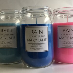 Puede incluir: Tres velas de vidrio en tarros con etiquetas que dicen "RAIN FOR AFTER THE MARY JANE RAIN FOREST SCENTED CANDLE HANDMADE IN MILWAUKEE WI CRUDE CANDLES". Las velas son azules, azul claro y rosas.