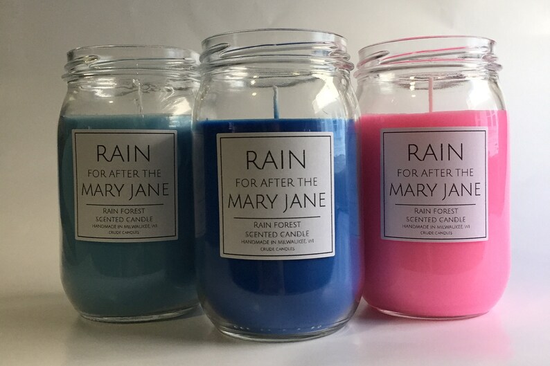 Puede incluir: Tres velas de vidrio en tarros con etiquetas que dicen "RAIN FOR AFTER THE MARY JANE RAIN FOREST SCENTED CANDLE HANDMADE IN MILWAUKEE WI CRUDE CANDLES". Las velas son azules, azul claro y rosas.