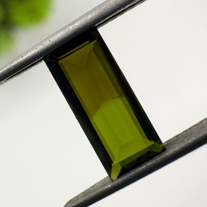 Könnte beinhalten: Ein rechteckiger, olivgrüner Edelstein, der von einer Metallpinzette gehalten wird. Der Edelstein hat einen facettierten Schliff, der seine Klarheit und Farbe zeigt. Der Edelstein ist ein leuchtendes Grün mit polierter Oberfläche.