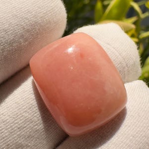 Puede incluir: Una gema pulida, de forma cuadrada, en un suave tono rosa melocotón. La piedra tiene bordes redondeados y una superficie lisa y brillante. La gema se sostiene en una tela blanca.