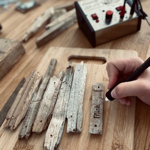 Puede incluir: Una persona utiliza una herramienta de pirograbado para grabar la palabra "Mint" en una pieza de madera. La madera es de color marr&oacute;n claro y tiene una textura rugosa. La herramienta de pirograbado es negra y tiene un bot&oacute;n rojo.