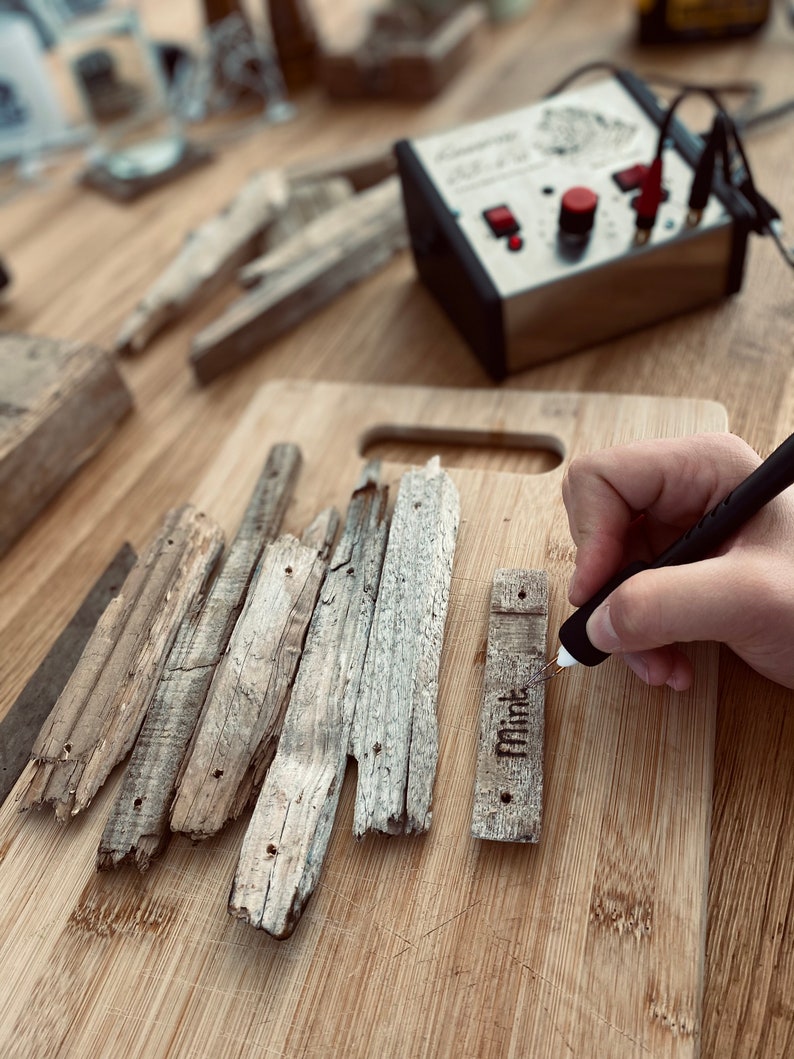 Puede incluir: Una persona utiliza una herramienta de pirograbado para grabar la palabra "Mint" en una pieza de madera. La madera es de color marr&oacute;n claro y tiene una textura rugosa. La herramienta de pirograbado es negra y tiene un bot&oacute;n rojo.