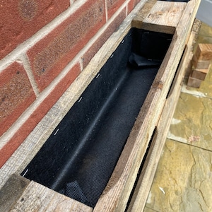 Puede incluir: Una jardinera de madera con tres compartimentos, cada uno forrado con tela negra. La jardinera est&aacute; montada en una pared de ladrillo.