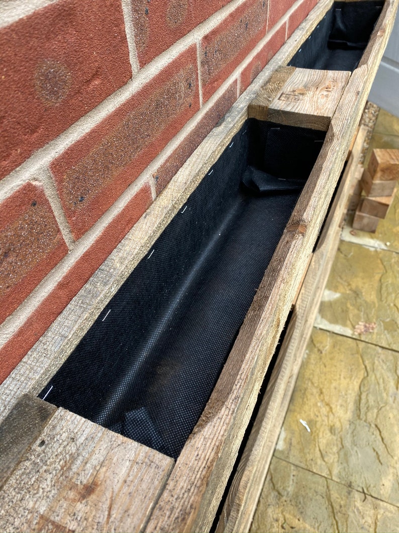 Puede incluir: Una jardinera de madera con tres compartimentos, cada uno forrado con tela negra. La jardinera est&aacute; montada en una pared de ladrillo.