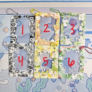 May include: Six decorative clear plastic frames with colorful embellishments and attached chains. Each frame has a different design, including stars, gears, and cute illustrations. Numbers 1 through 6 are written in red on each frame.