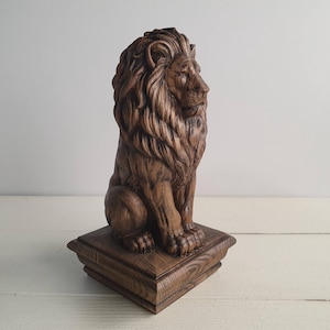 May include: A brown wooden lion sculpture featuring a detailed mane and facial features, depicted in a seated position. The lion is mounted on a square, tiered wooden base with a visible wood grain texture throughout the entire piece.