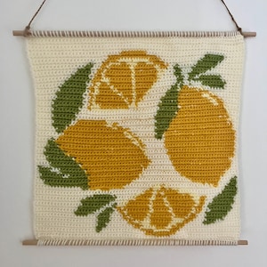 Peut inclure: Une tenture murale au crochet avec un motif de citron. L'œuvre présente trois citrons jaunes et des feuilles vertes sur un fond blanc cassé. Elle est suspendue par une cheville en bois et une ficelle marron.