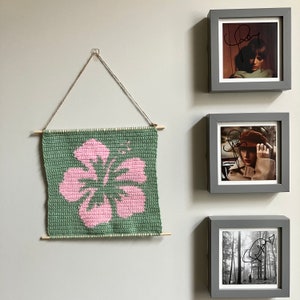 Peut inclure: Une tapisserie murale en crochet vert avec un motif de fleur d'hibiscus rose. La tapisserie est exposée sur un mur blanc avec trois images encadrées.