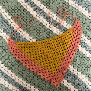 May include: A crocheted triangle scarf with a mustard yellow center and a pink border. The scarf is lying on a green and white crocheted blanket.