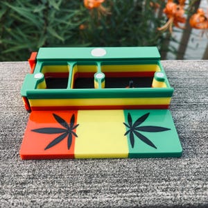 Puede incluir: Un recipiente rectangular multicolor con tapa abatible. El recipiente es principalmente verde, amarillo y rojo, con una base a juego con diseños de hojas de cannabis. El diseño recuerda la bandera rastafari.