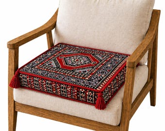 Puede incluir: Cojín cuadrado con un detallado patrón geométrico en rojo, negro y gris, colocado sobre una silla beige. El cojín tiene borlas rojas en las esquinas. La silla tiene brazos de madera y un cojín beige. Mide aproximadamente 40 cm x 40 cm.