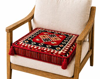 Puede incluir: Un cojín cuadrado con un patrón geométrico rojo, negro y blanco descansa sobre una silla beige. El cojín tiene borlas rojas en las esquinas y un borde a rayas. La silla tiene brazos de madera y un cojín beige.