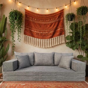 Puede incluir: Un sofá modular gris con cuatro cojines grises y un reposabrazos a juego. Un tapiz de pared tejido de color óxido con flecos color crema cuelga sobre el sofá. La habitación está decorada con plantas colgantes y una alfombra roja estampada.
