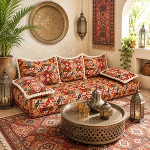 May include: A low-profile floor sofa featuring a vibrant, multi-colored geometric pattern with white tassel trim. The set includes three large back cushions and two smaller side cushions, paired with a round, etched metal coffee table.