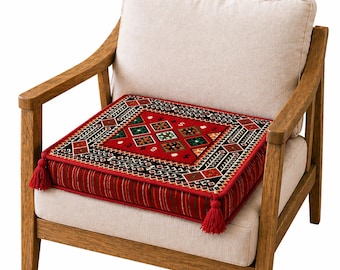 Puede incluir: Un cojín cuadrado rojo con un patrón geométrico en negro, verde y blanco descansa sobre una silla beige. El cojín tiene borlas rojas en las esquinas y un borde a rayas. La silla tiene brazos de madera y un asiento y respaldo beige.