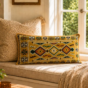 May include: A rectangular cushion with a yellow base and a geometric pattern in green, orange, and white. The cushion has a yellow border and is placed on a window seat next to a textured beige cushion.