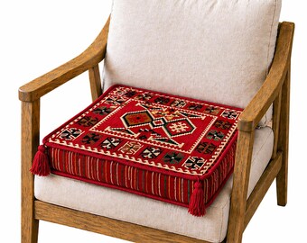 Puede incluir: Un cojín cuadrado rojo con un patrón geométrico en negro, marrón y crema está sobre una silla beige. El cojín tiene borlas rojas en cada esquina y un borde a rayas. La silla tiene brazos y patas de madera.