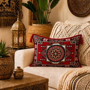 May include: A rectangular throw pillow featuring a vibrant red base with a central geometric mandala pattern in black, white, and teal. The pillow is accented with small red tassels at each corner and has a woven texture throughout the fabric.