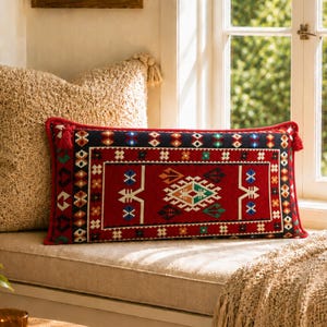 Pode incluir: Uma almofada retangular decorativa com um centro vermelho com um desenho geométrico em branco, verde, azul e laranja. A almofada tem uma borda azul escura com padrões coloridos e borlas vermelhas em cada extremidade. Descansa sobre um parapeito bege.
