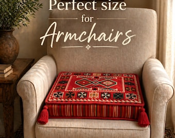 Puede incluir: Un cojín rojo y negro estampado con borlas sobre un sillón beige. El cojín tiene un diseño geométrico y el texto "Perfect size for Armchairs". La habitación tiene un ambiente cálido.