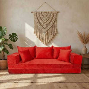 May include: A vibrant red velvet sofa with matching throw pillows. A woven macrame wall hanging and potted plant add to the room's decor. The sofa sits on a neutral-toned rug, creating a cozy living space.