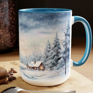 May include: A large white ceramic mug with a blue interior and handle. The mug features a winter landscape scene with a cabin, snow-covered trees, and a snowy sky. A spoon and a macaron are on the wooden surface.