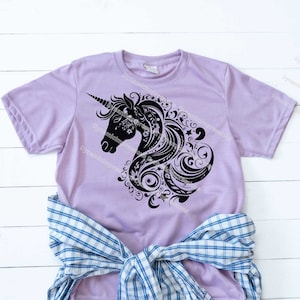 以下が含まれることがあります： ラベンダー色のTシャツに、黒いシルエットで描かれた花柄のユニコーンの頭部がデザインされています。