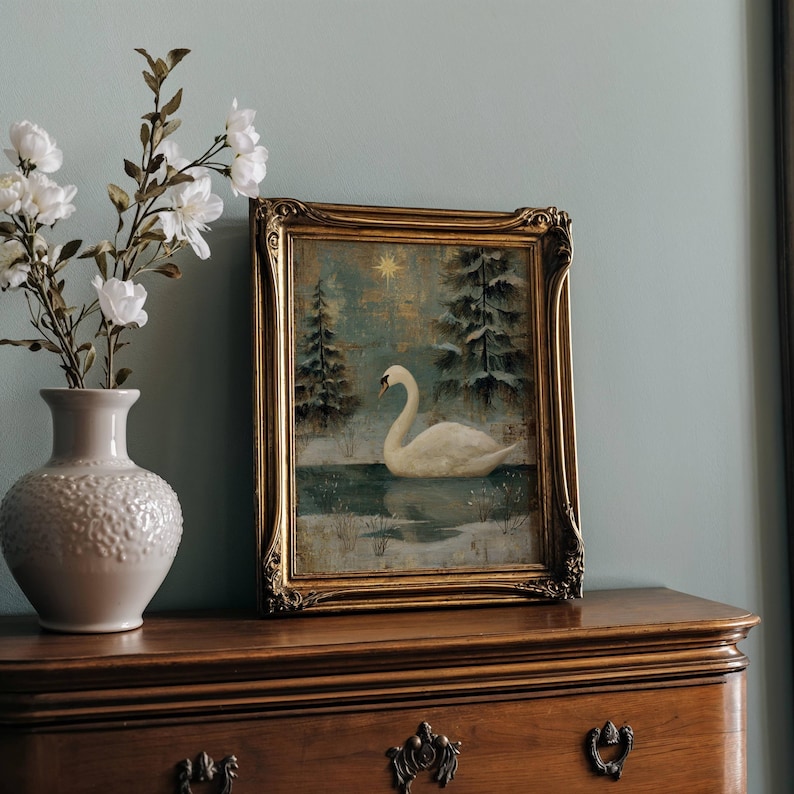 May include: A framed painting of a swan on a lake, with snow-covered trees and a star in the background. The painting is in a gold ornate frame, displayed on a wooden dresser. A white vase with white flowers is to the left.