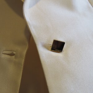 Op de afbeelding: Een close-up van een cr&egrave;mekleurige blazer met een vierkante gouden knoop waarop "VERSACE" staat.