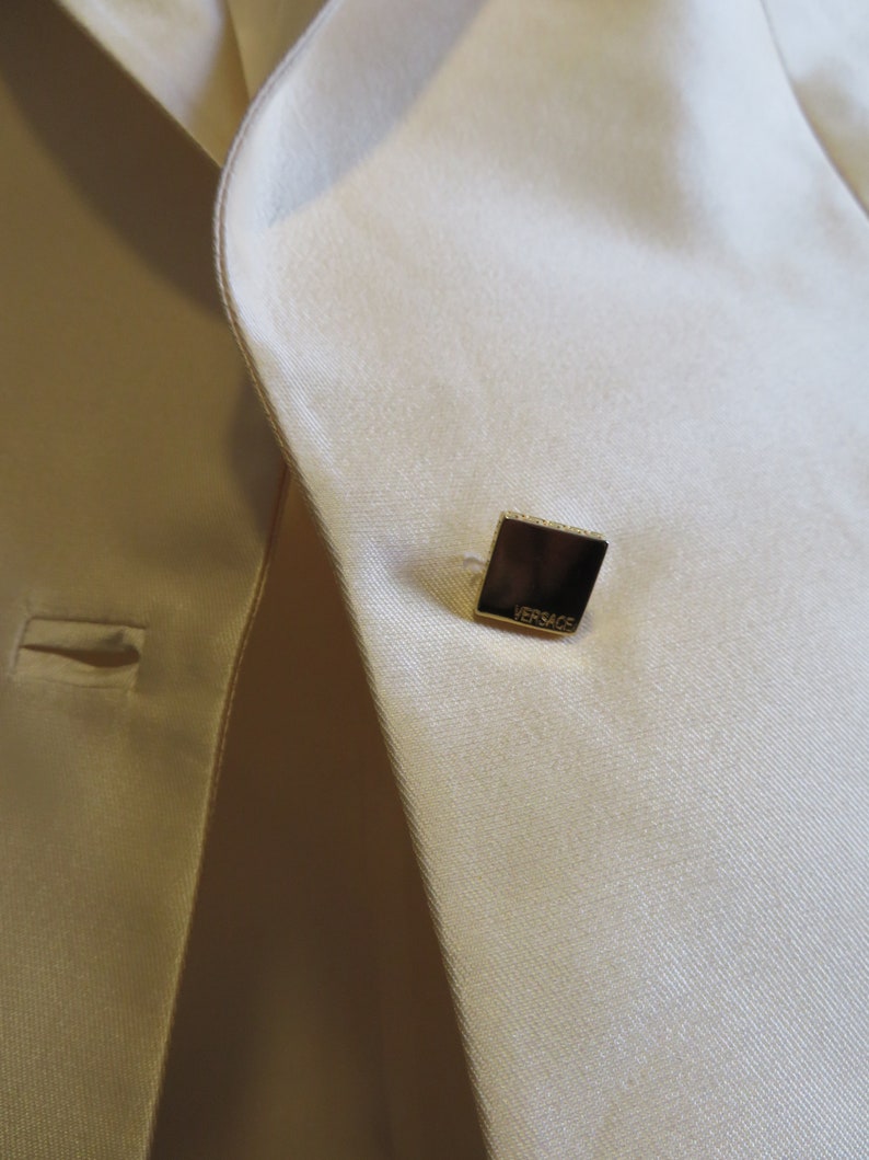 Op de afbeelding: Een close-up van een cr&egrave;mekleurige blazer met een vierkante gouden knoop waarop "VERSACE" staat.