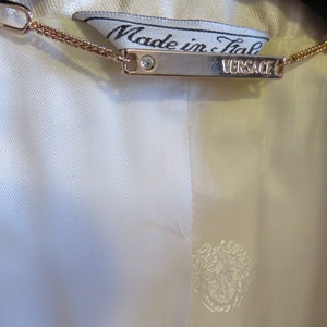 Op de afbeelding: Een wit label met de tekst "Made in Italy" en een zilveren tag met de tekst "VERSACE" bevestigd aan een gouden ketting.