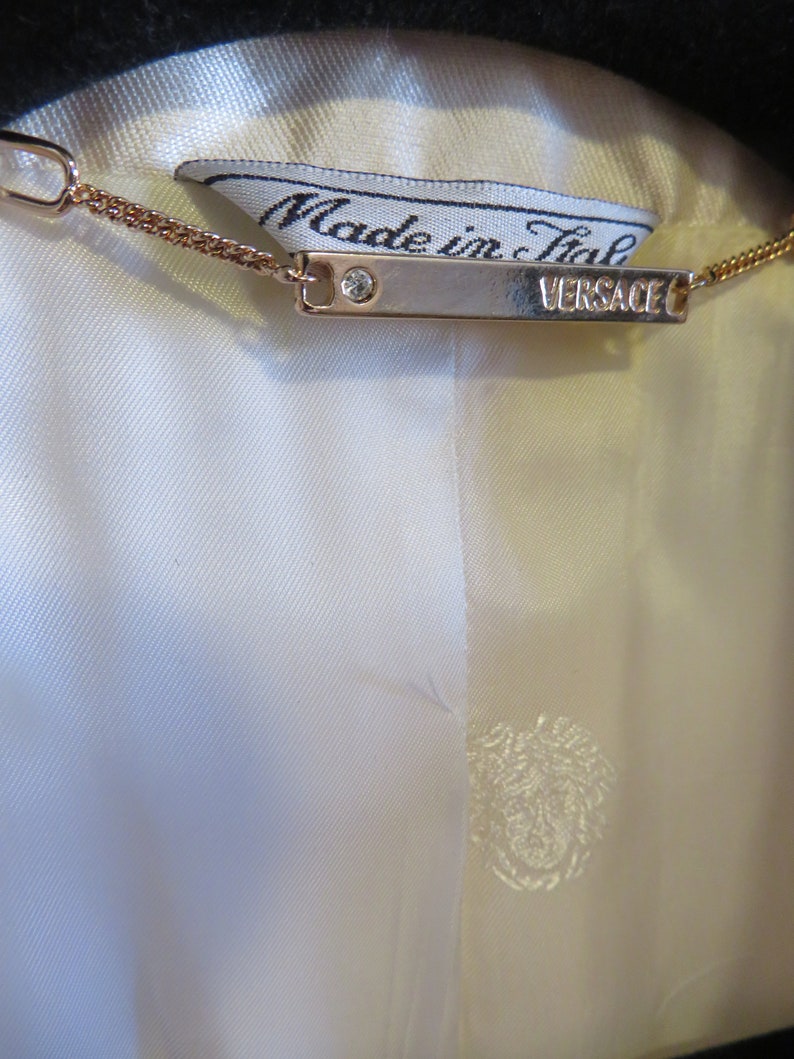 Op de afbeelding: Een wit label met de tekst "Made in Italy" en een zilveren tag met de tekst "VERSACE" bevestigd aan een gouden ketting.