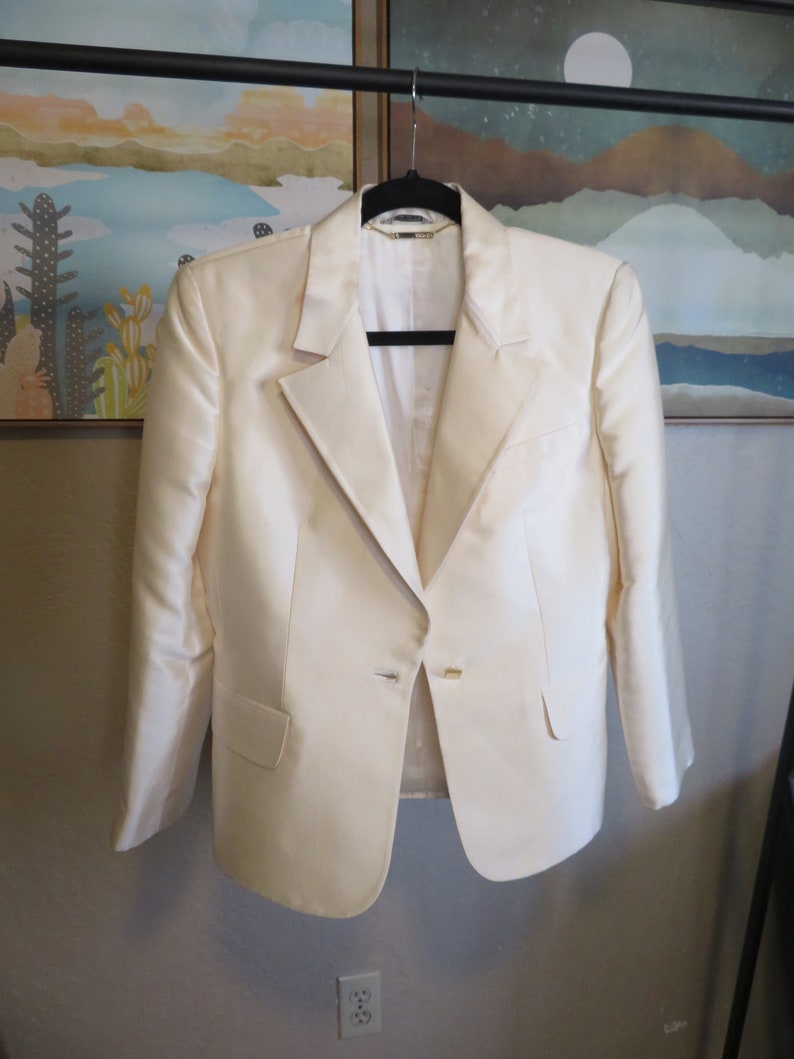 Op de afbeelding: Een wit blazer met een enkele knoopsluiting en een gouden knoop. De blazer hangt aan een zwarte hanger.