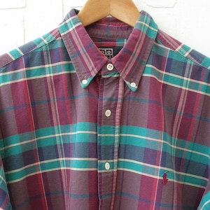 May include: A long-sleeved button-down shirt featuring a plaid pattern in shades of plum, teal, and cream. The shirt has a button-down collar, a front button placket with white buttons, and a small embroidered logo on the lower left chest area.