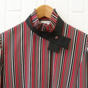 May include: A striped long-sleeved blouse with a black collar and a black bow tie. The blouse has red, gray, black, and white stripes.