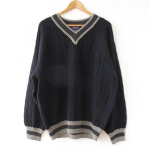 May include: A navy blue cable-knit sweater with a V-neck and gray striped trim on the collar, cuffs, and hem. The sweater is hanging on a wooden hanger against a white background. The brand name "GANT" is visible on a small tag.