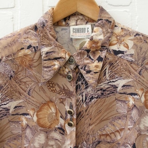 May include: A long-sleeve button-up shirt in shades of tan, brown, and black with an all-over botanical print featuring leaves and flowers. The shirt has a pointed collar and a label at the neckline that reads CONRAD C. COLLECTION MADE IN CANADA.