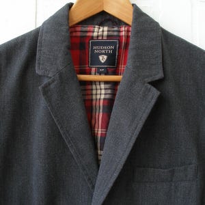 May include: A dark grey blazer with a notched lapel and a single pocket. The interior lining has a red and black plaid pattern. The label reads "HUDSON NORTH". The blazer is hanging on a wooden hanger.