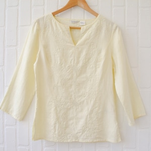 May include: A pale yellow long-sleeve blouse featuring a split V-neckline and tonal floral embroidery detailing on the front panel. The garment has a straight hemline and is displayed on a wooden hanger against a white brick-patterned background.